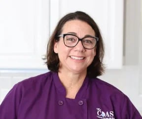 Woman wearing glasses and purple CAAS branded scrubs, smiling at camera in a clinical setting